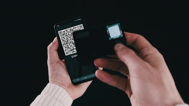 Young man uses portable cold crypto wallet to scan QR code on a smartphone for a transaction. Using modern device camera for sending Bitcoin on black background. Digital cash, safe, future concept. 4K
