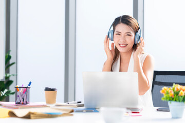 Naklejka premium Asian businesswoman wearing headphones to listen to music in the office to relieve stress after work Listening to music made her happy and smiling.