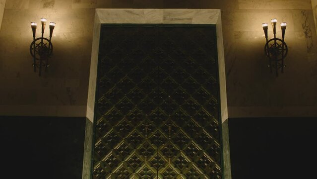 Huge metal medieval castle door in Dark place . Old vintage chandeliers , lamps or lanterns on the wall . Big tall antique door inside church , cathedral , palace or house . Camera moving on door