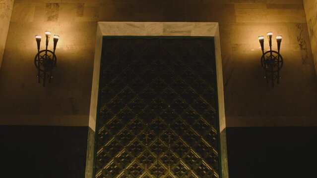 Huge metal medieval castle door in Dark place . Old vintage chandeliers , lamps or lanterns on the wall . Big tall antique door inside church , cathedral , palace or house . Camera moving on door