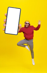 Fototapeta Young man in Uk flag sunglasses jumping in joy like ballet dancer holding smartphone wearing casual red hoodie and jeans isolated on yellow background. Mobile app advertising product placement obraz