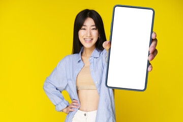 Asian girl holding smartphone showing white screen, and excited smile on camera wearing blue shirt isolated on yellow background. Great offer mobile app advertisement. Product placement