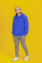 Young man model in casual wear, glasses posing looking at camera on yellow background with hands in pockets. Stylish bearded smart hipster man casual look. Thoughtful guy on yellow background