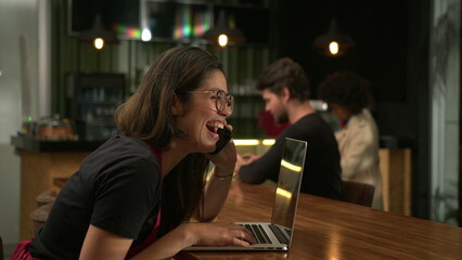 Happy girl staff talking on phone smiling and laughing browsing online orders sitting at coffee shop table