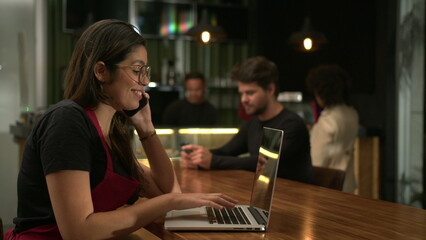 Barista receiving orders on phone browsing online with laptop at cafe. Small business concept of staff using computer at coffee shop