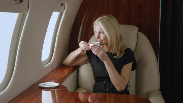Beautiful successful mature business woman travels on private plane. Portrait of rich senior woman drinking tea or coffeee while enjoying luxury journey in first business class airlines