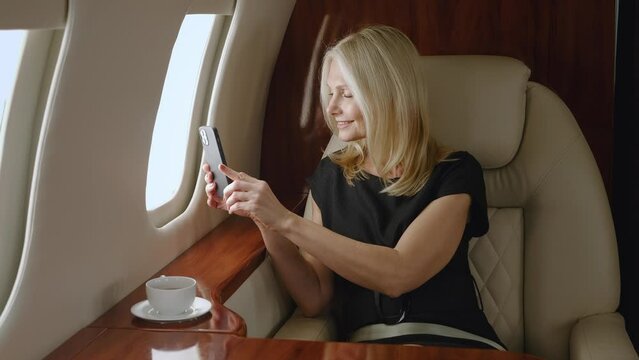 Beautiful mature woman millionaire taking photos with smartphone from window of aircraft. Businesswoman traveling in private jet or first business class. Luxury travel journey.