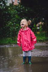 Beautiful girl walking along the puddles under the summer rain in a water-repellent raincoat and boots, on a green background of trees and grass. Carefree childhood. Happy child.