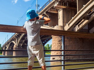 Unrecognizable nine-year-old caucasian boy weared casual clothes photographs a bridge over a river. View from the back.