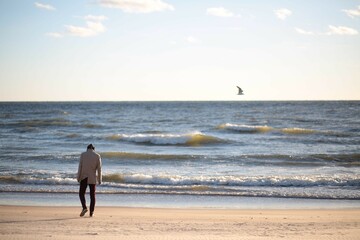 A man in a raincoat and a hood walks along a sandy beach in windy weather at sea