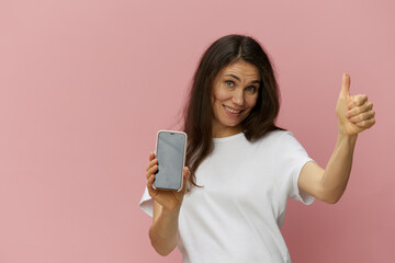 Fototapeta premium a beautiful, happy woman with long dark hair stands in a light t-shirt with a phone in her hands and shows a thumbs up