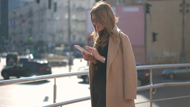 Side view beautiful millennial woman in sunglasses and coat standing on city street surfing social media on smartphone. Absorbed Caucasian young lady in sunshine outdoors in urban town