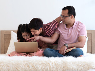 Fototapeta premium Indian father's hand pointing to the laptop for two teenage children ,son and daughter, sitting on the bed together at home. Family using laptop for movie, homework, online learning.