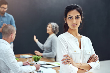 Its time to get serious about our business. Portrait of a businesswoman standing with her arms folded in the boardroom while a colleague gives a presentation in the background.