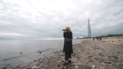 Woman takes photos on camera of sea in cloudy weather. Action. Professional photographer woman takes pictures of sea standing on background of cloudy sky