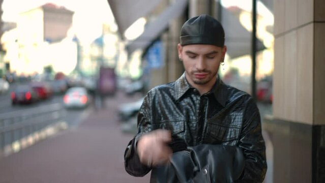 Front view portrait of chilling young gay man strolling on city street putting on leather jacket leaving. Confident Caucasian handsome LGBTQ person walking in urban city outdoors. Lifestyle concept