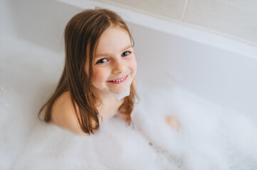A small, smiling, beautiful red-haired girl with long hair, the child bathes, washes in a white bath with soap foam and hot water. Happy childhood. - obrazy, fototapety, plakaty