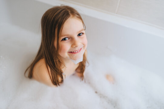 A small, smiling, beautiful red-haired girl with long hair, the child bathes, washes in a white bath with soap foam and hot water. Happy childhood.