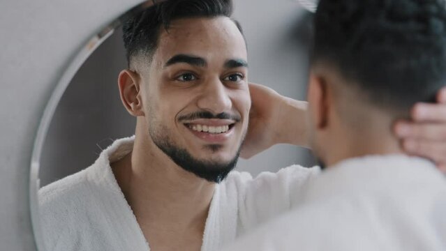 Mirror reflection headshot Indian Arabian bearded male macho bearded guy touching hair after showering enjoy new hairstyle shampoo smiling standing in bathroom in morning in hotel bath in white robe 