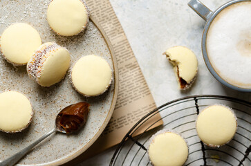 Cornstarch alfajores with milk caramel and coconut on a beige plate and on a black rack, and a cup of latte on light grey background.