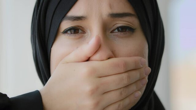 Female portrait indoors Islamic Arabian girl sad afraid Muslim woman Arab lady in black hijab looking at camera covers mouth with hands quiet silence discrimination stop talking oppression of women