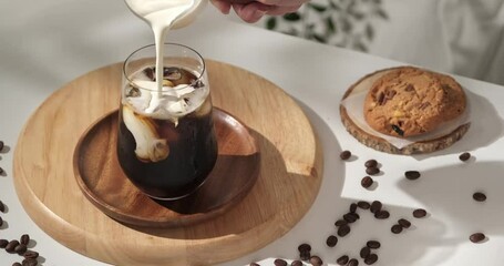 4k video, pouring creamy milk into a brewing iced coffee is served with cookies. Cold coffee drink glass with ice and cream milk on white table. in slow motion. - Powered by Adobe