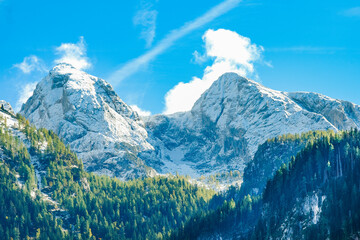 View of the mountain peaks covered with snow, in the foreground mountains overgrown with coniferous forest. Sunny day, blue sky, clouds.