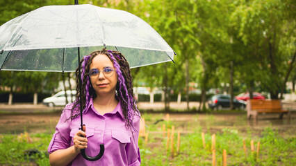 Happy woman with umbrella. Cheerful female in a purple shirt holding umbrella and looking at camera while standing in green park on rainy day