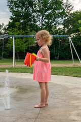 Little child playing with water and toys at splash pad in the local public park during hot summer day. Small beautiful girl in pink dress standing at fountain playground.