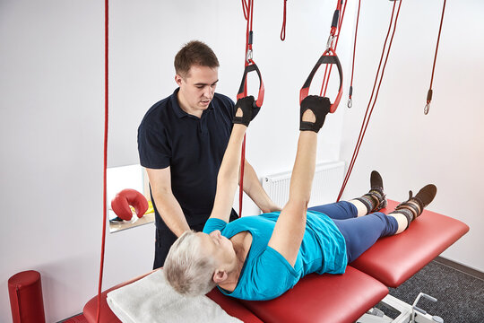 Rehabilitation specialist doing active treatment on suspension straps to elderly patient. Therapeutic exercises to restore pain-free movement patterns and improve function on red cord slings
