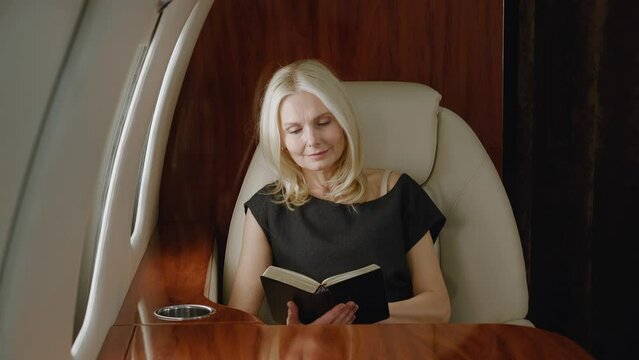 Beautiful mature rich woman reading book or notebook while flying in private airplane. Luxury traveling in private jet.