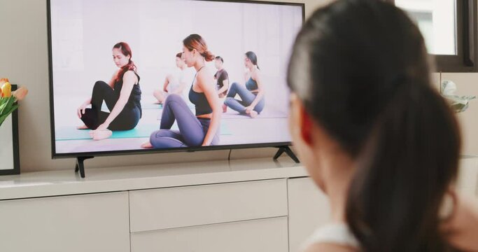Young attractive Asian woman in sportswear site on mat in the floor virtual fitness class with group of people on a video conference on television practice yoga for beginner in living room at home.