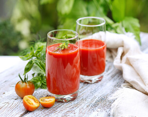 Fresh tomato juice in glass glasses, with tomato slices on a light wooden table in the garden against the backdrop of greenery