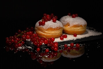 Berlinere German sweet donuts sprinkled with powdered sugar filled with fruit jam surrounded by ripe berries on a black slate board on a black background with reflection