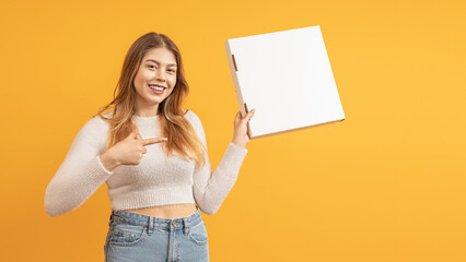 Fototapeta na wymiar smiling young woman holding a pizza box in one hand and pointing her finger at it on a colored background with space for text