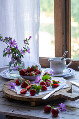 Tagliere con fragole sparse tagliate a metà e intere e fiori di malva di fronte a una finestra