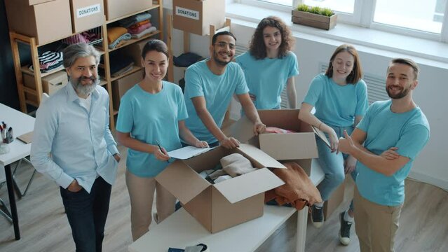 Portrait of happy people volunteers standing in charity company office smiling showing thumbs-up hand gesture and looking at camera. Youth and humanitarian aid concept.
