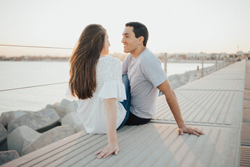  Hispanic man and a smiling brunette girl are sitting on a breakwater in Spain