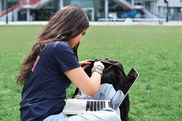 Young girl student sitting on green grass lawn on campus with laptop computer studying