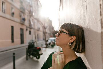 Close up profile portrait of short haired stylish girl in sunglasses closed eyes and gently smiling in sunlight near the wall. Portrait of woman in black shirt on street