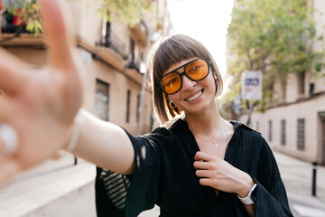 Charming smiling girl with short hairstyle in black blouse and yellow sunglasses is making selfie...