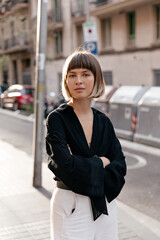 Fototapeta premium Outside portrait of stylish pretty European girl with straight short hairstyle wearing black blouse and white pants posing at camera in sunlight on summer street 