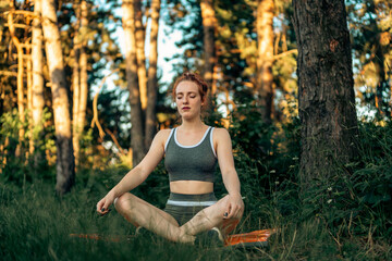 A young fit woman practices meditation on a yoga mat against the backdrop of a forest lit by the setting sun. Exercise and training outdoors. Healthy way of living. A sporty woman doing yoga in nature