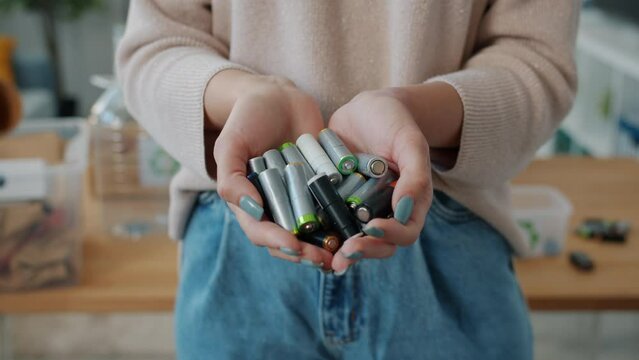 Close-up of female hands holding batteries with recycling boxes visible in background in apartment. Ecological problems and zero waste concept.