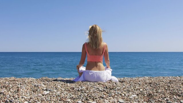 Adult blonde woman meditating on the pebble beach facing the sea