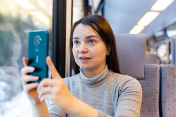 Interested female traveler sitting near window on comfortable seat in express train, filming...