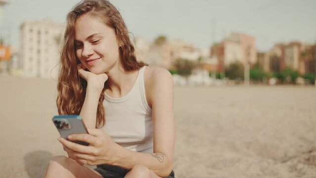Smiling girl with mobile phone in her hands sits on the sand on the beach on modern building background. Beautiful girl is watching videos and photos on her smartphone