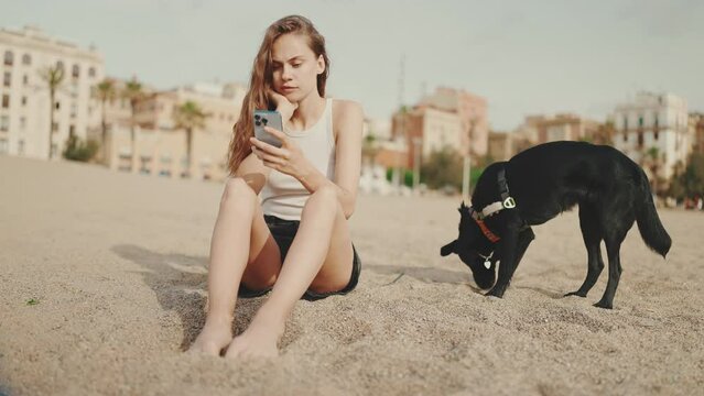 Cute girl with mobile phone in her hands sits on the sand on the beach with black dog on modern building background. Beautiful girl is watching videos and photos on her smartphone