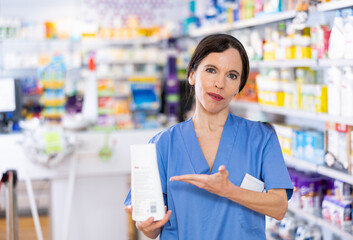 Friendly young adult woman pharmacist standing at drugstore with medical product in hands