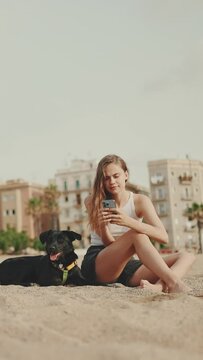 VERTICAL VIDEO: Cute girl with mobile phone in her hands sits on the beach with black dog on modern building background. Girl is watching videos and photos on her smartphone